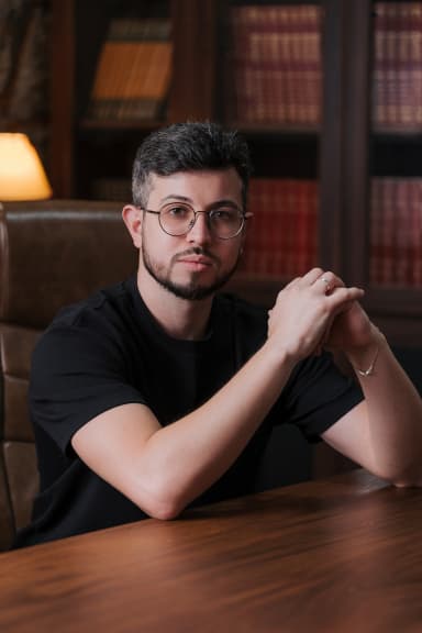 Portrait of Gabriel Tsunoda: man with round glasses, beard and black t-shirt, sitting at a wooden table with hands clasped, in front of a bookshelf under warm lighting